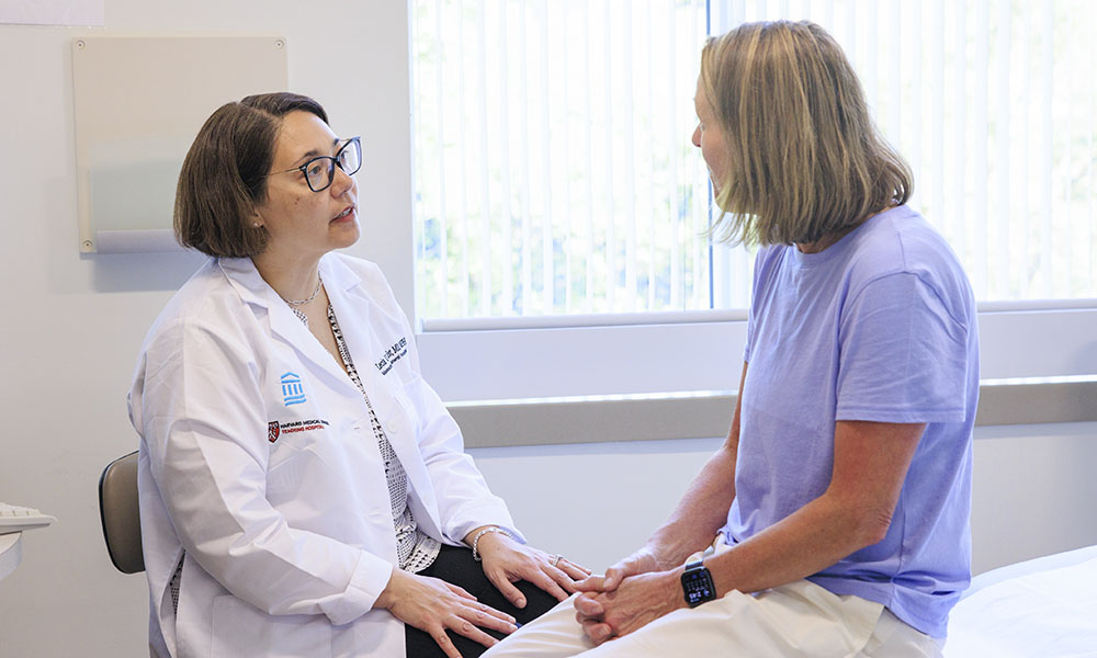A clinician in a white coat sits and speaks with a patient in an exam room. A clinician in a white coat sits and speaks with a patient in an exam room.